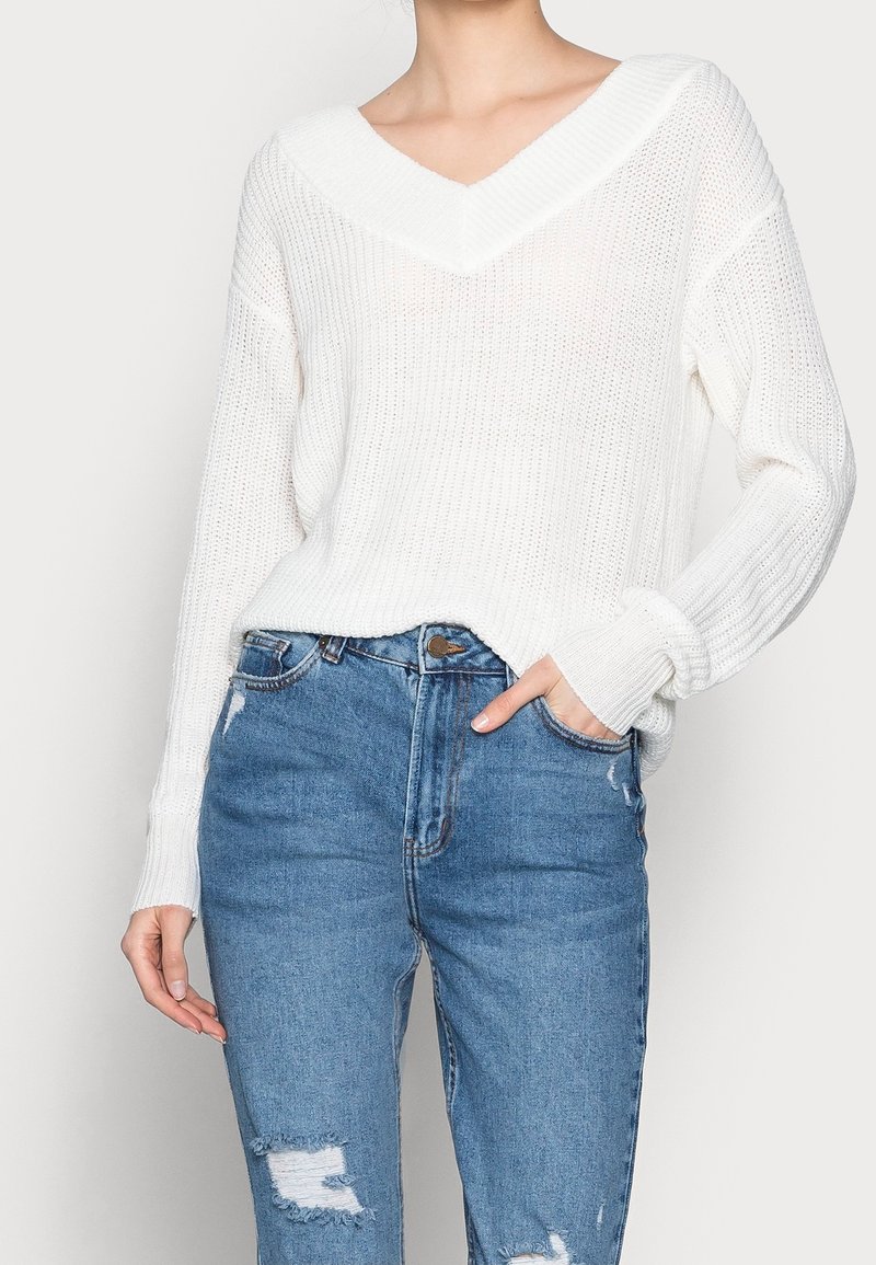Femme portant un pull en maille blanc à col en V et un jean bleu déchiré, une main dans la poche et l'autre détendue sur le côté.