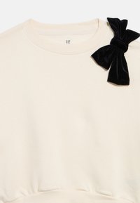 Krémovo sfarbená detská mikina s okrúhlym výstrihom a čiernym zamatovým mašľou pripevnenou na pravom ramene.