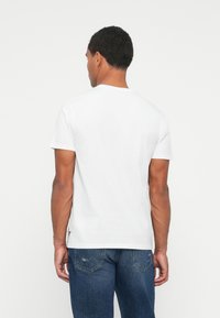 Mand, der står med ryggen til, iført en almindelig hvid t-shirt med korte ærmer og blå denim jeans foran en ensfarvet hvid baggrund.
