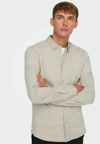 Camisa de manga larga beige claro con cuello abotonado, textura suave y un brillo sutil. Presenta cierre con botones y dobladillo redondeado.