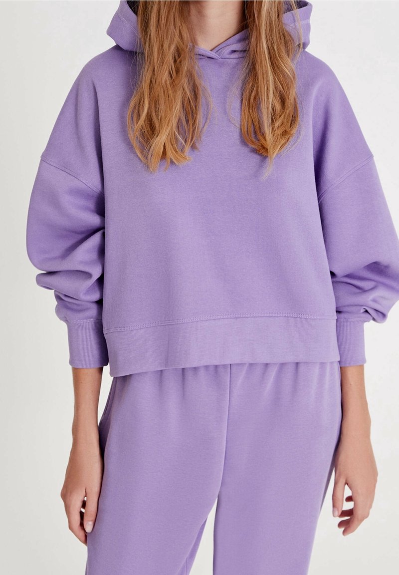 Sweat à capuche lavande ample et pantalon de jogging assorti, porté par une personne aux longs cheveux ondulés, visible du cou jusqu'aux hanches.