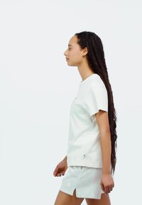 Weißes kurzärmeliges T-Shirt mit rundem Ausschnitt, kombiniert mit hellgrauen Shorts mit seitlichen Schlitzen und Logo am Saum. Lange, strukturierte Haare.