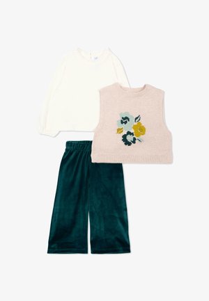 Chemise blanche à manches longues, gilet en tricot sans manches beige avec un motif de fleurs bleues et jaunes, et pantalon large en velours vert foncé.