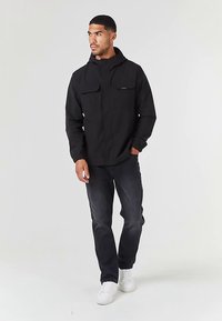 Juoda striukė su gobtuvu ir dviem priekinėmis kišenėmis su atvartais, derinama su tamsiais džinsais ir baltais sportbačiais. Audinys atrodo lygus ir universalus.