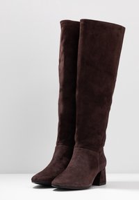 Botas de ante marrón por encima de la rodilla con punta afilada y tacón cuadrado, que presentan una textura lisa y detalles de costura mínimos.