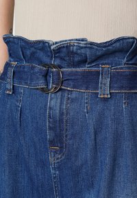 Pantalones de mezclilla azul de talle alto con una cinturilla fruncida, que cuentan con una hebilla circular de metal oscuro y sutiles detalles de costura naranja.