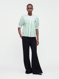 Femme debout, une main dans la poche, portant un chemisier vert rayé à manches bouffantes et un pantalon noir à jambes larges.