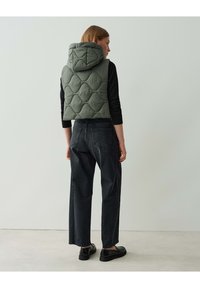 Gepolsterte olivgrüne Steppweste mit Kapuze, getragen über einem schwarzen Langarmoberteil. Schwarze hochgeschnittene Röhrenjeans und schwarze Schuhe.