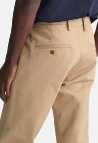 Chinos en coton beige avec une texture lisse, dotés d'une poche arrière fermée par un bouton marron et d'une coupe ajustée. Coutures visibles en accent.