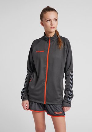 Junge Frau trägt eine dunkelgraue Sportjacke und Shorts mit orangem Rand und Hummel-Logos, steht und schaut nach rechts.