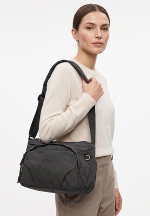Femme en pull beige et pantalon portant un sac à bandoulière noir GMV avec bandoulière réglable sur fond blanc.