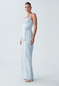 Jasnoniebieski kombinezon z dopasowaną górą, regulowanymi ramiączkami i wiązaniem w talii. Wykonany z miękkiego materiału o gładkiej fakturze.