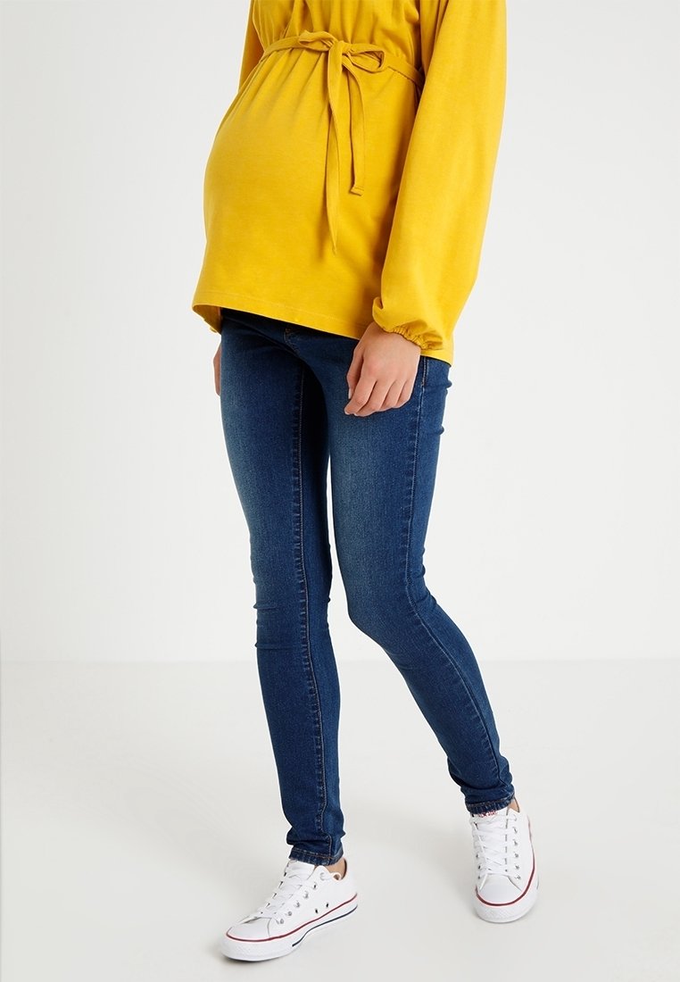 Graviditetstopp i ljusgul tyg, med knytning i midjan, kombinerad med blå skinny jeans och vita sneakers. Slät textur och figurnära design.