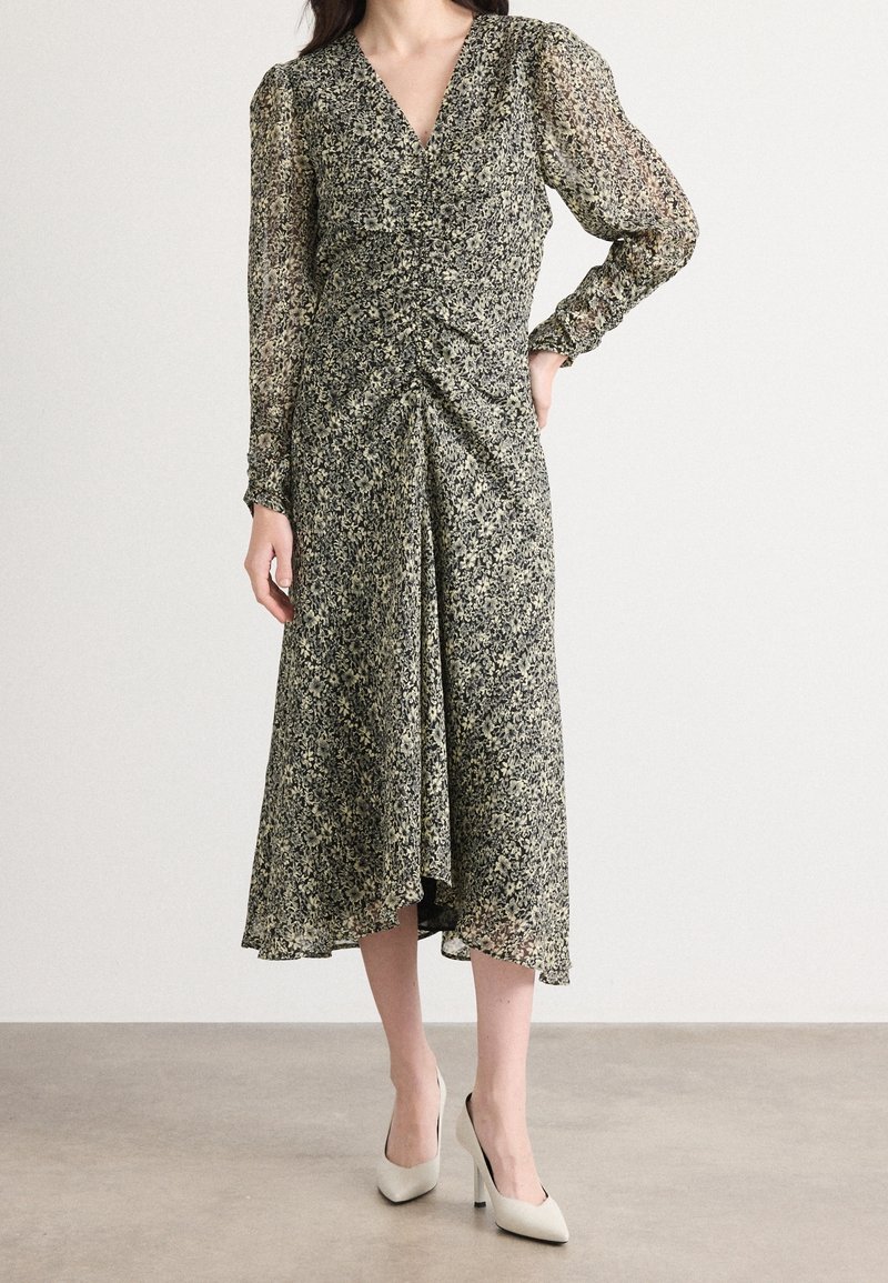 Robe midi à imprimé floral en noir et crème avec un col en V, des manches longues en voile transparent bouffantes, et un détail froncé à la taille. Associée à des talons blancs.