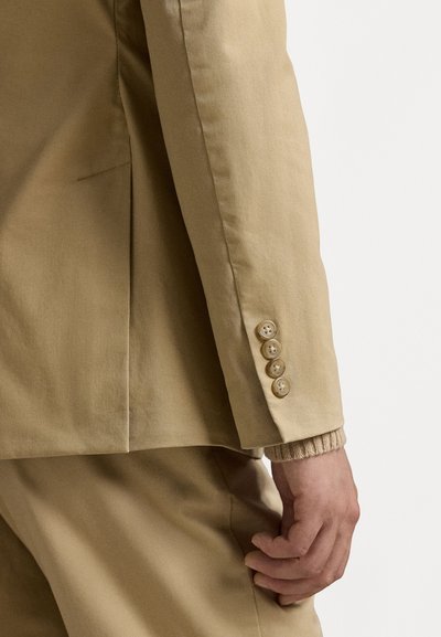 Béžový kalený blazer s jemnou texturou, s pěti knoflíkovými detaily na rukávu a diskrétním skladem na zádech.