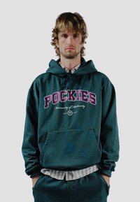 Sudadera de color verde oscuro con "POCKIES" en letras burdeos en negrita. Presenta un bolsillo delantero, capucha con cordón y un ajuste informal. Tejido texturizado.