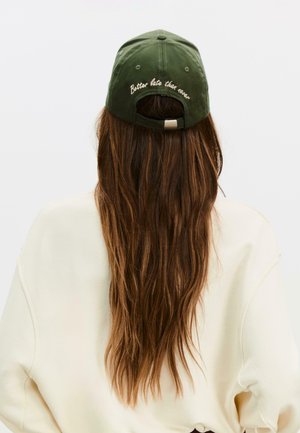 Cappellino da baseball in cotone verde, con cinturino posteriore regolabile, ricamato con la scritta "Meglio tardi che mai" in marrone chiaro sul pannello posteriore.