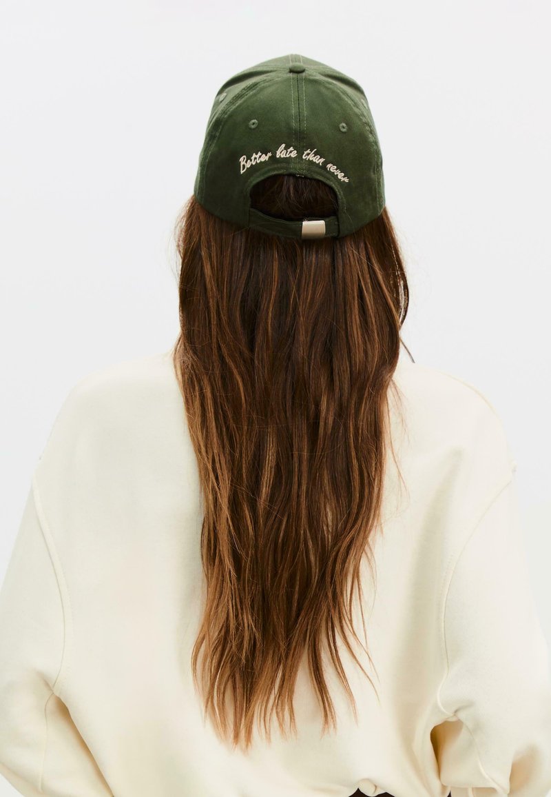 Cappellino da baseball in cotone verde, con cinturino posteriore regolabile, ricamato con la scritta "Meglio tardi che mai" in marrone chiaro sul pannello posteriore.