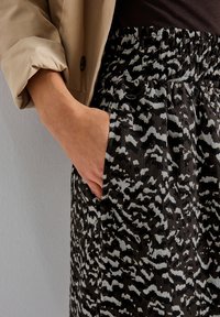 Main dans la poche d'une jupe taille haute à motif abstrait noir, blanc et gris, à côté de la manche de veste beige retroussée.