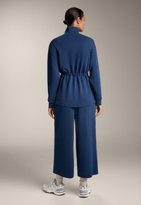 Temno modra, lahka bluzica z visokim ovratnikom, elastičnim pasom; v kombinaciji z širokimi hlačami. Bele superge z modrimi akcenti dopolnjujejo obleko.