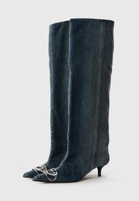 Botas hasta la rodilla en mezclilla oscura con un diseño ajustado, punta puntiaguda y un lazo plateado como acento en la zona de la punta. Acabado de tela texturizada.