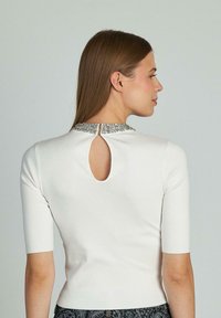 Top bianco aderente con maniche corte, caratterizzato da una schiena a chiave e un colletto impreziosito. Texture liscia con una leggera lucentezza.