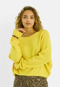 Mujer con cabello rizado que lleva un suéter amarillo brillante y pantalones con estampado de leopardo, posando con una mano cerca de su rostro y una ligera sonrisa.