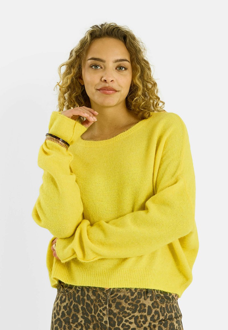 Mujer con cabello rizado que lleva un suéter amarillo brillante y pantalones con estampado de leopardo, posando con una mano cerca de su rostro y una ligera sonrisa.