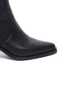 Černá kožená kotníková bota s špičatou špičkou, hladkým povrchem, minimálním prošíváním a nízkou, plochou gumovou podrážkou.