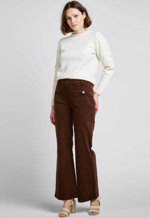 Pantalons en velours côtelé marron évasés avec poches avant et fermeture à bouton, associés à un pull texturé blanc et des sandales à talons beiges.