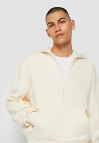 Hoodie zippé de couleur crème avec capuche à cordon, présentant une texture lisse et des poches avant. Porté par-dessus un t-shirt blanc.