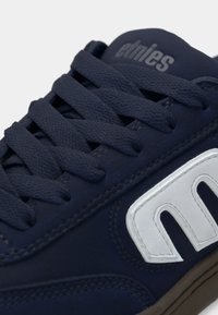 Ναυτικό μπλε αθλητικό παπούτσι με υφή από σουέτ υλικό, διαθέτοντας επίπεδα κορδόνια, λευκή λογότυπο λεπτομέρεια και επενδεδυμένη γλώσσα. Διακριτή ραφή ορατή.