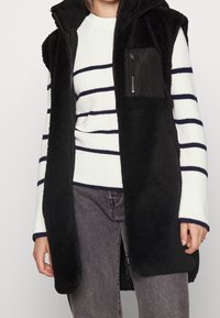 Czarna kamizelka z polaru z kapturem, z kieszenią na piersi na zamek, noszona na białym i granatowym swetrze w paski, zestawiona z szarymi dżinsami.