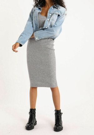 Mujer con vestido midi gris acanalado, chaqueta vaquera corta azul claro y botas negras de combate con cordones, de pie con los brazos parcialmente cruzados.