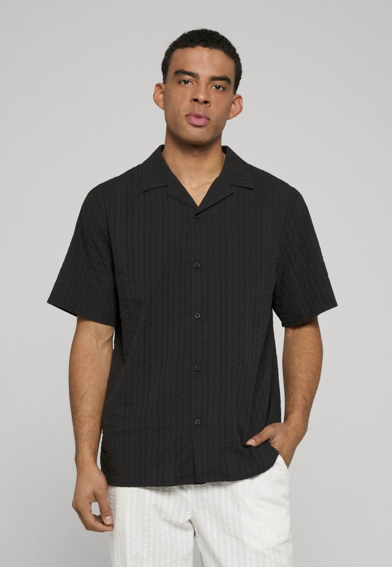 Chemise noire à manches courtes avec une texture côtelée verticale, fermeture à boutons et col plat. Portée avec un pantalon blanc à motifs.