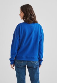 Blue cotton sweatshirt with a relaxed fit, ribbed cuffs, and hem. Worn over blue jeans, showing a simple design without distinctive patterns.