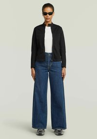 Giacca nera di cotone con collo alto, camicia bianca sotto, abbinata a jeans larghi blu e sneaker grigie. Occhiali da sole scuri completano il look.