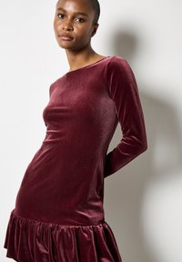 Burgundowa aksamitna sukienka z długimi rękawami, dopasowanym gorsetem i falbanowym wykończeniem, prezentująca gładką teksturę i subtelny połysk.