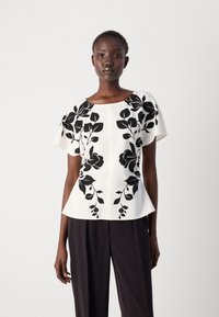 Witte top met zwarte bloemenpatronen, korte mouwen, een bootnek en een uitlopende silhouet, gecombineerd met donkere broeken.