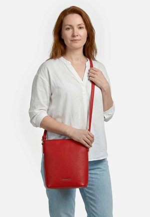 Mujer con cabello rojo que lleva una blusa blanca y jeans azules, sosteniendo un bolso bandolera de cuero rojo con acabado texturizado.