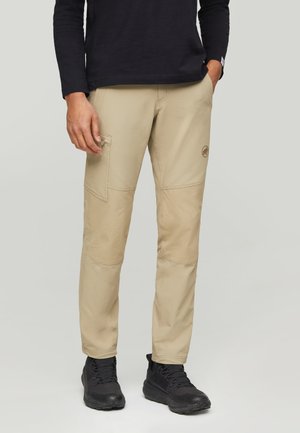 Mann trägt beige Cargohose mit Seitentaschen, schwarzes Langarmshirt und schwarze Wanderschuhe, steht mit einer Hand in der Tasche.