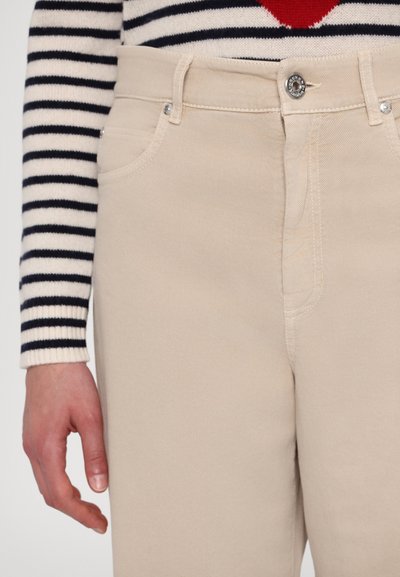 Beige katoenen broek met een aansluitend ontwerp, voorzien van zijzakken en subtiele stikdetails. Draagt een gestreepte trui.