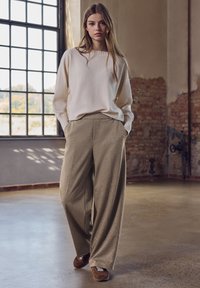 Sweter z dzianiny w kolorze kremowym z prążkowanymi mankietami zestawiony z szerokimi, beżowymi spodniami o fakturowanej powierzchni. Modelka ma na sobie brązowe sneakersy i stoi w przestronnym wnętrzu.