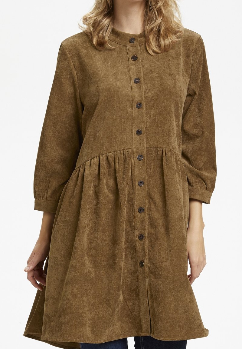 Femme portant une robe ample en velours côtelé marron à manches longues et boutons foncés devant, debout devant un fond uni.