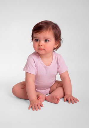Egy barna hajú kisgyerek keresztbe tett lábakkal ül, világos rózsaszín, rövid ujjú rugdalózót visel, fehér háttér előtt, kissé oldalra nézve.