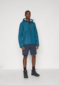 Τιρκουάζ αδιάβροχο μπουφάν με φερμουάρ και κουκούλα, συνδυασμένο με σκούρες βερμούδες. Το σύνολο περιλαμβάνει μαύρες ορειβατικές μπότες και έχει ανακλώσες λεπτομέρειες.