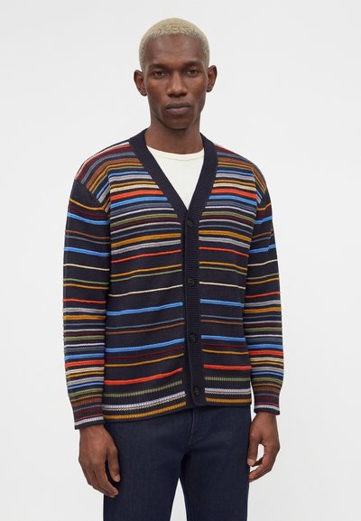 Άνδρας που φοράει πολύχρωμη οριζόντια ριγέ ζακέτα πάνω από λευκό πουκάμισο και σκούρο παντελόνι, στέκεται σε απλό φόντο.