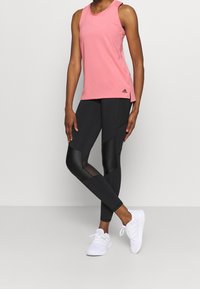 Personne portant un débardeur rose, un legging de sport noir avec des panneaux en mesh et brillants, et des baskets blanches, debout devant un fond uni.