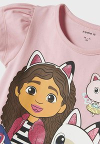 Camiseta rosa de manga corta con un personaje de dibujos animados de pelo rizado castaño, que lleva una diadema brillante y detalles gráficos coloridos y juguetones.
