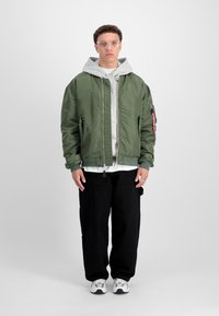 Veste bomber vert olive avec une fermeture éclair à l'avant, des poignets côtelés et un écusson sur la manche, superposée à un sweat à capuche gris, associée à un pantalon cargo noir.